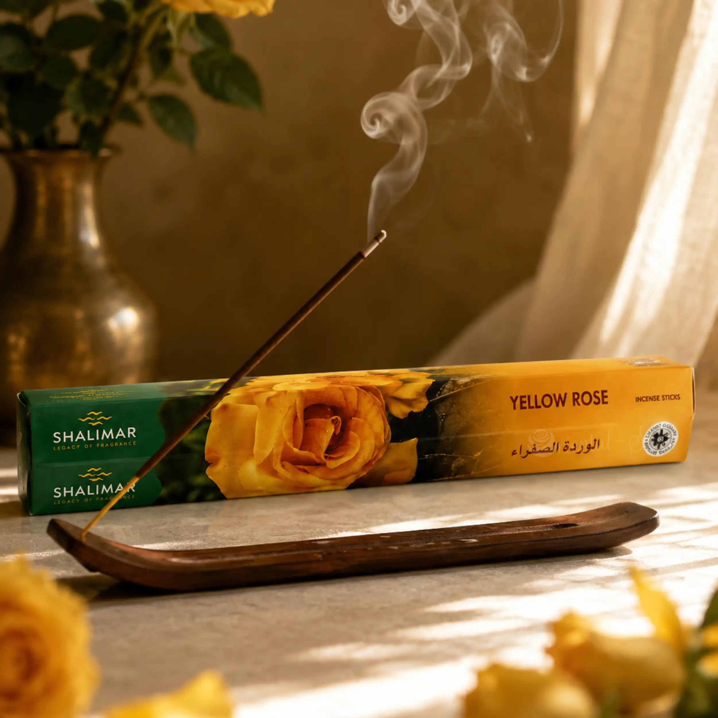 Brennendes Shalimar Yellow Rose Räucherstäbchen im Holzhalter mit sanftem Rauch und gelber Rosenverpackung im Hintergrund