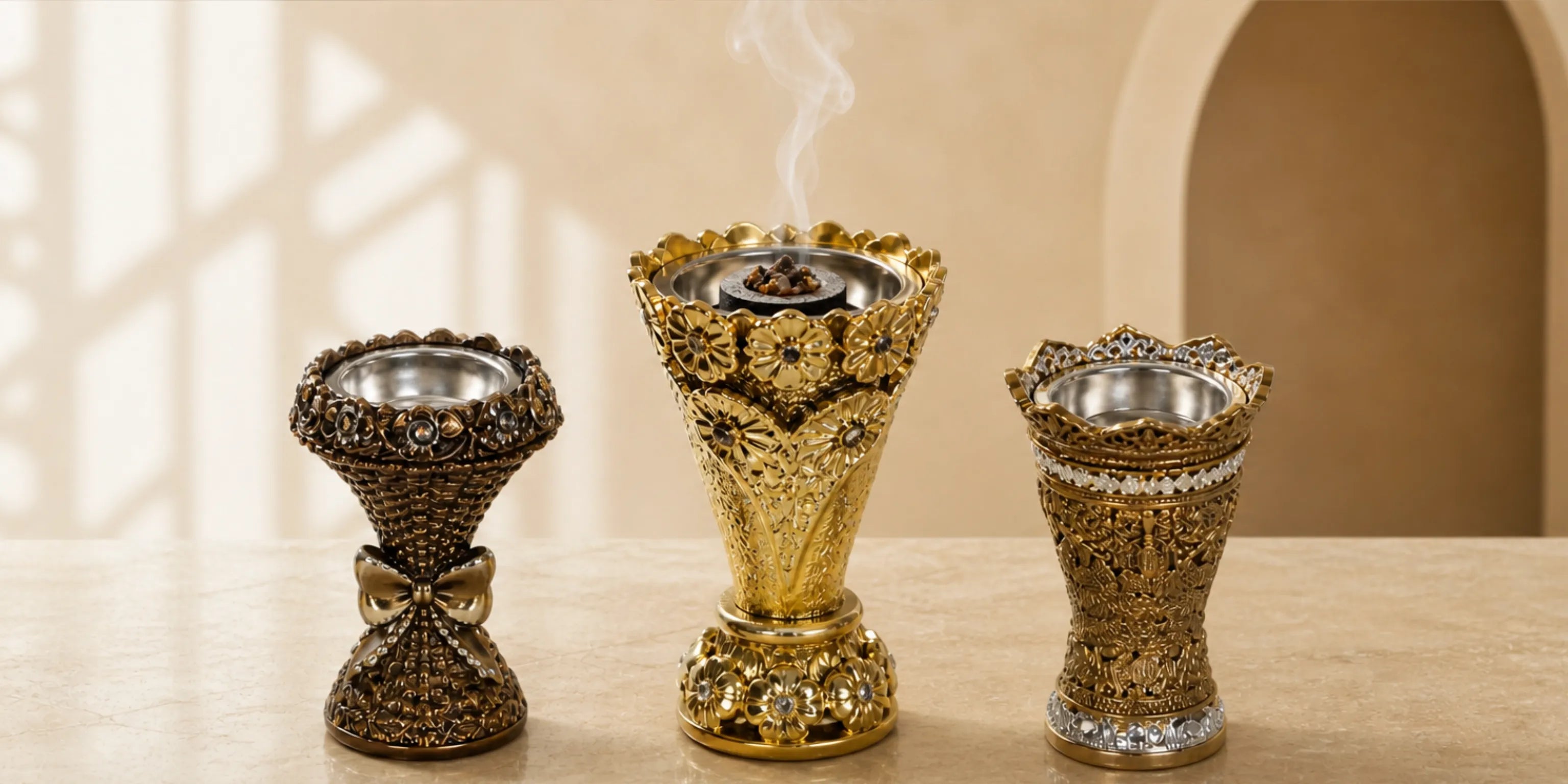 akhoorhalter Collection – drei offene goldene und bronzefarbene Mubkhara-Räuchergefäße mit Metallschalen und aufsteigendem Bakhoor-Duft­rauch