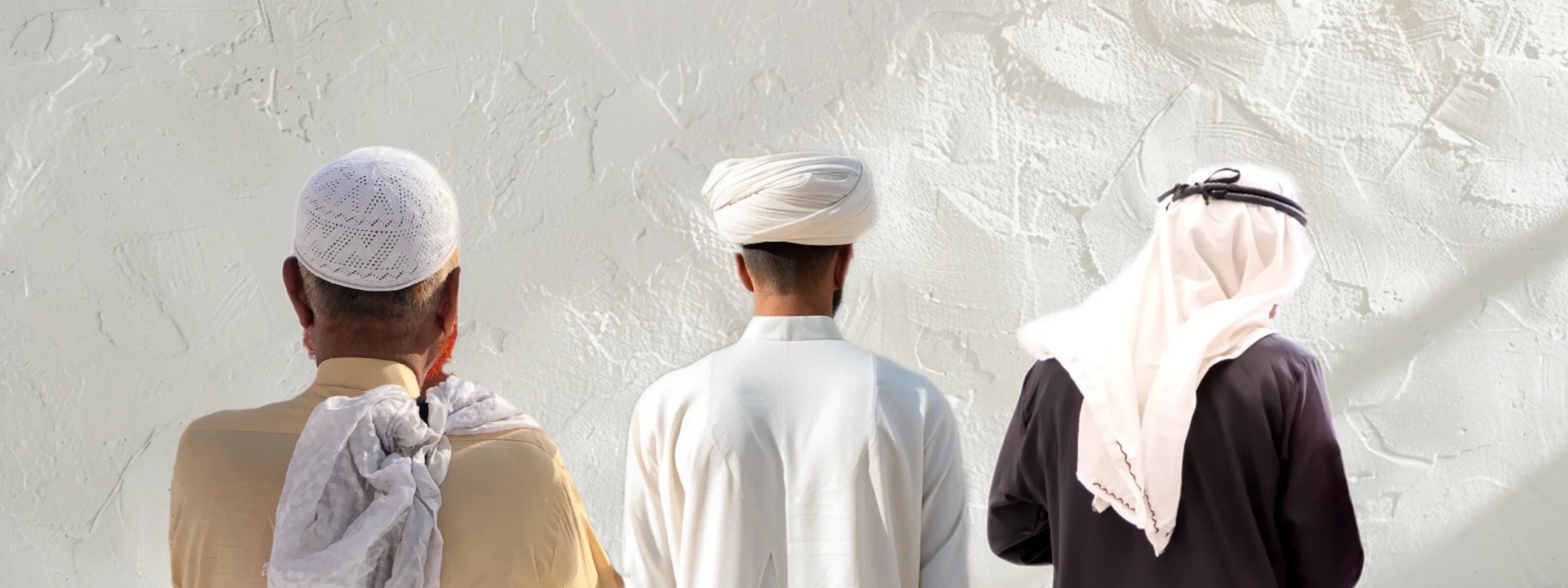 Drei Männer von hinten mit unterschiedlicher islamischer Kopfbedeckung – Takke, Turban und Shemagh – vor heller Wand